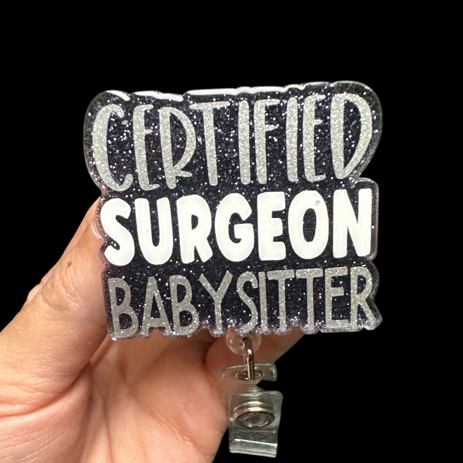 A hand holding a badge reel with the text 'CERTIFIED SURGEON BABYSITTER' on it.