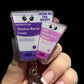 A hand holding a retractable badge reel with two badge toppers. Each topper has a playful design; one is labeled 'Moisture Barrier Cream' and the other 'Show Me The Booty' with a smiley face.
