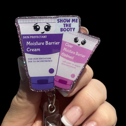 A hand holding a retractable badge reel with two badge toppers. Each topper has a playful design; one is labeled 'Moisture Barrier Cream' and the other 'Show Me The Booty' with a smiley face.
