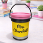 Yellow bucket with pink lid labeled 'on a desk with classroom items in the background