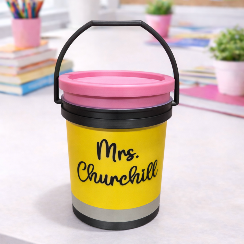 Yellow bucket with pink lid labeled 'on a desk with classroom items in the background